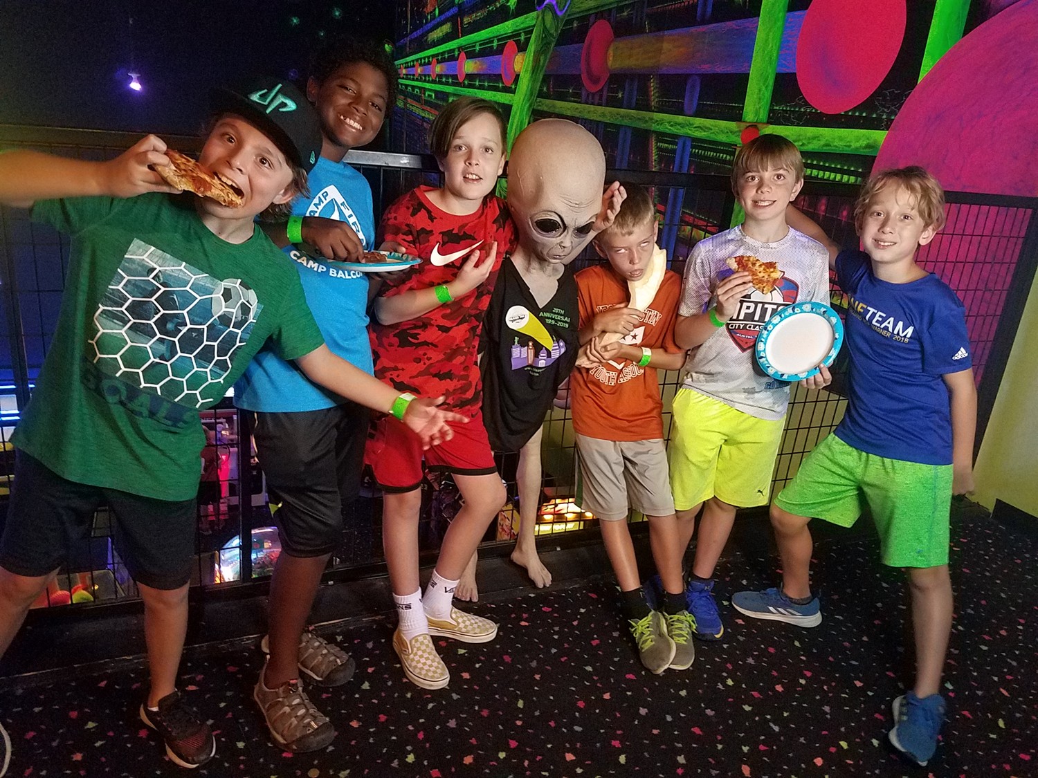 Kids posing with alien at arcade, holding pizza.